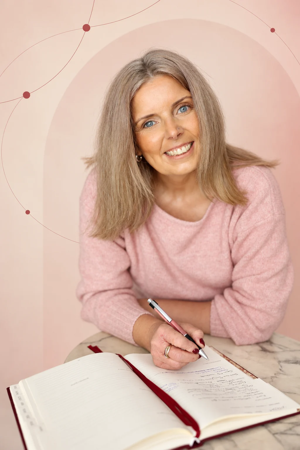 Emma, founder of Emmagraphy, smiling while writing in her notebook, against a soft pink branded background.